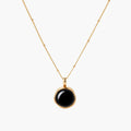 Circular black agate pendant on gold-plated sterling silver chain against cream background, showing modern minimalist design.