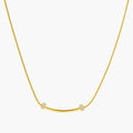 Delicate gold-plated necklace with curved bar pendant and crystal accents on snake chain against cream background.
