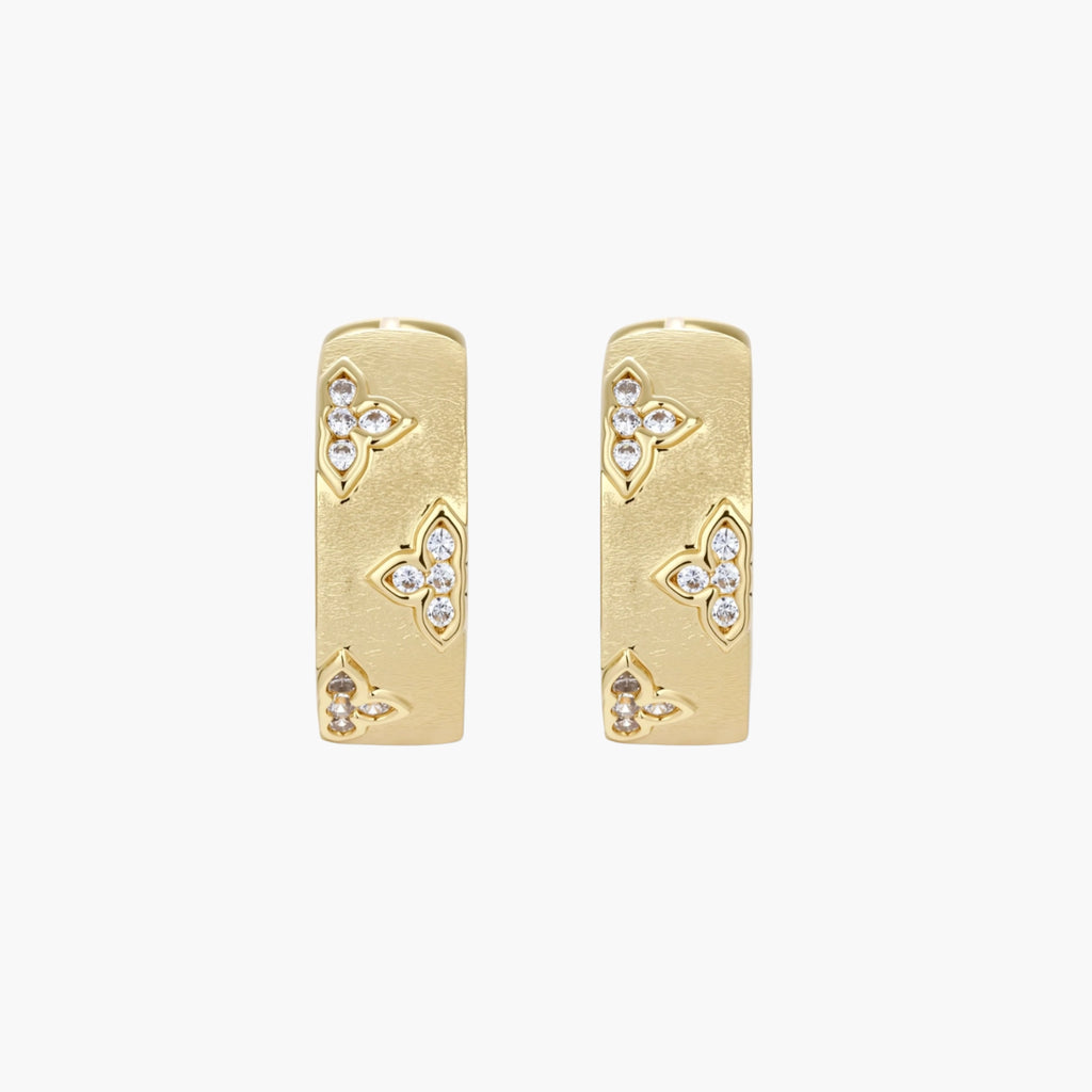 Gold-plated hoop earrings with textured surface and raised floral motifs set with clear zirconia stones on cream background.