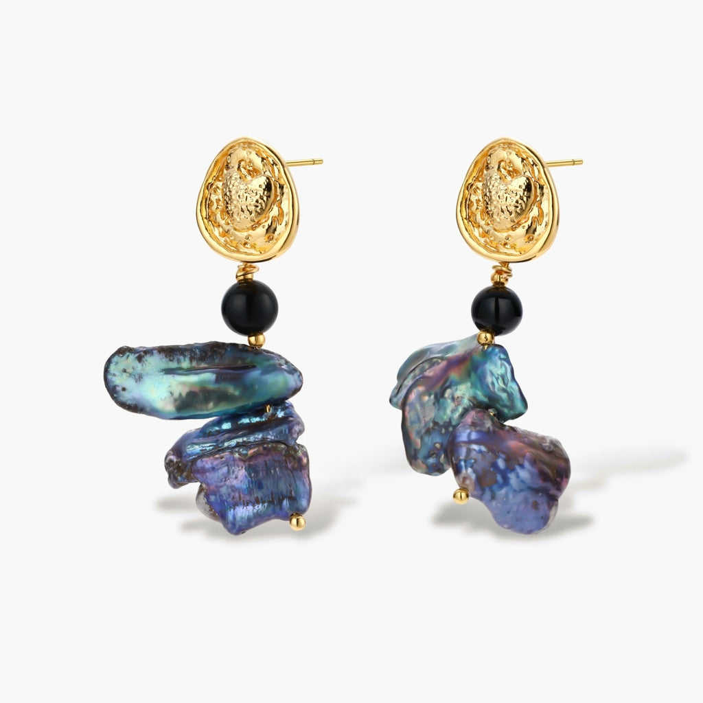 Ornate gold-plated drop earrings with black beads and iridescent blue-purple baroque pearls against white background.
